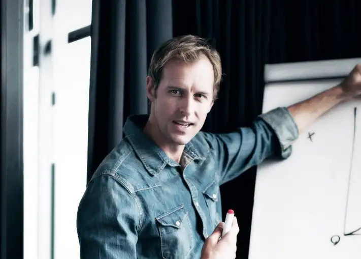 Young man in denim shirt leans forward, holding a red marker near a whiteboard.