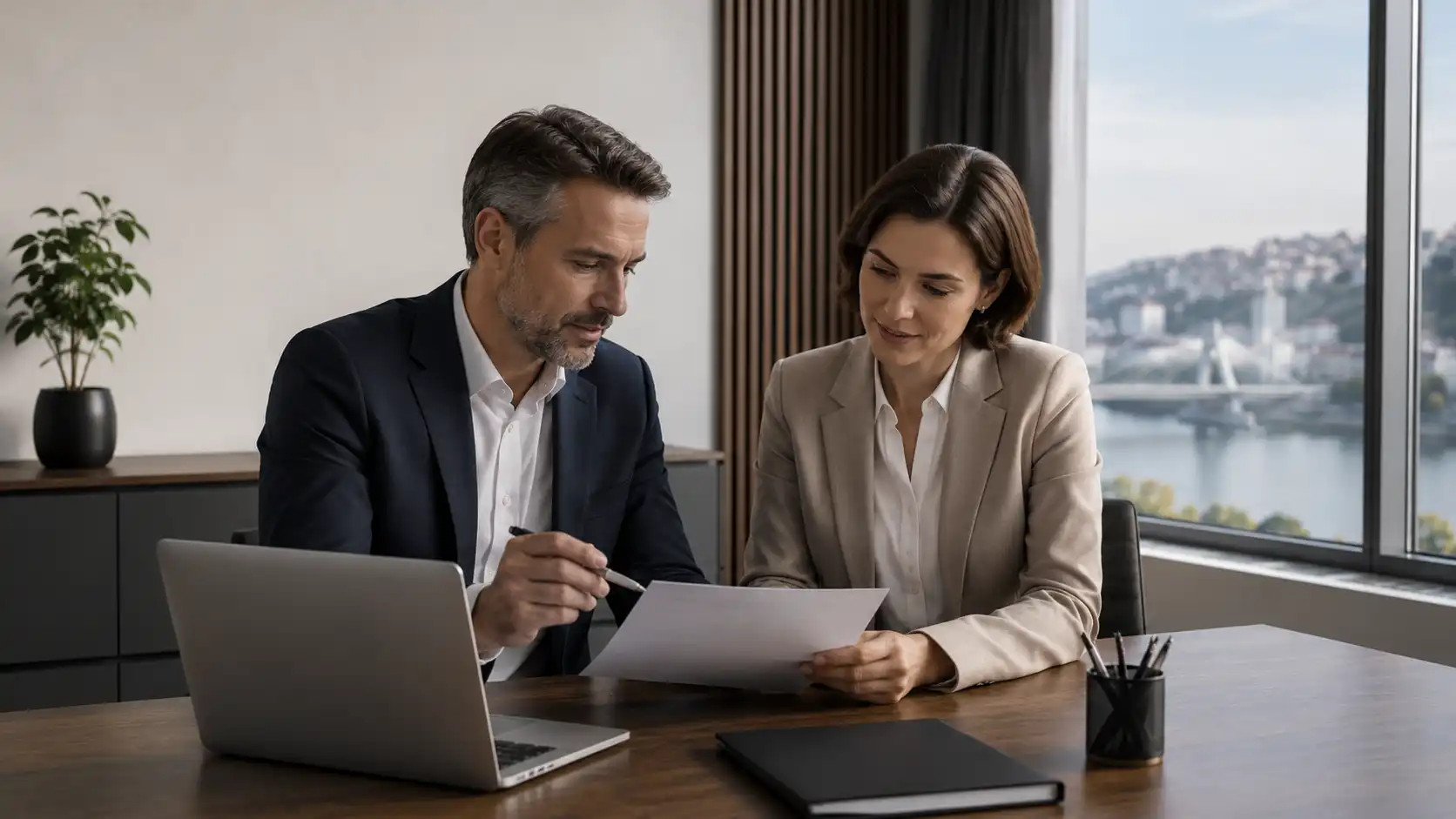 A man and a woman are sitting at a laptop in an office, looking through documents together by a large window with a view.