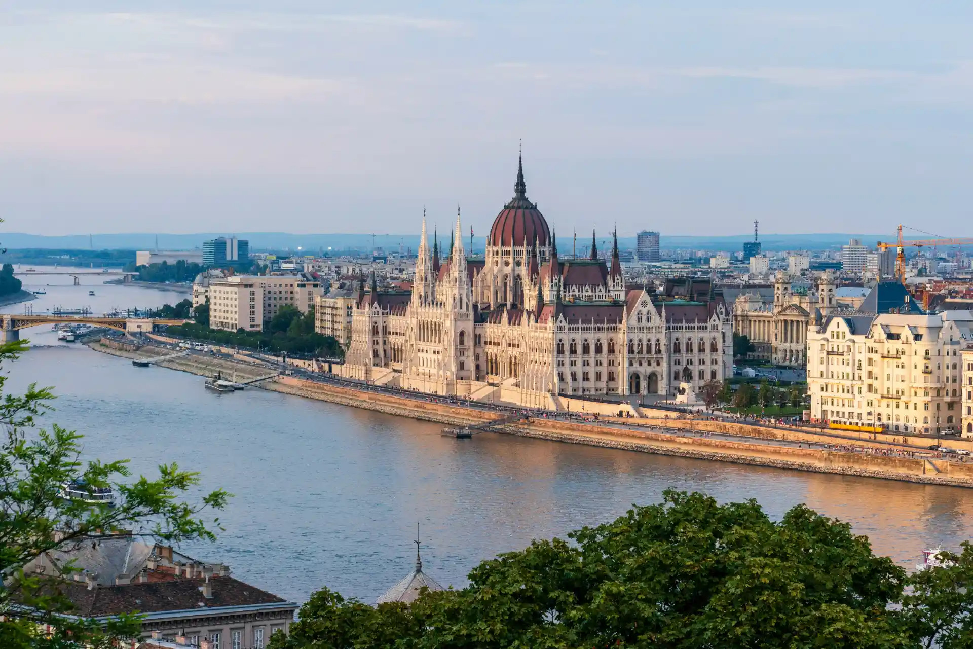 Pohľad na budapeštiansky parlament pri Dunaji, s vežami, nábrežím a mestskou panorámou v pozadí.
