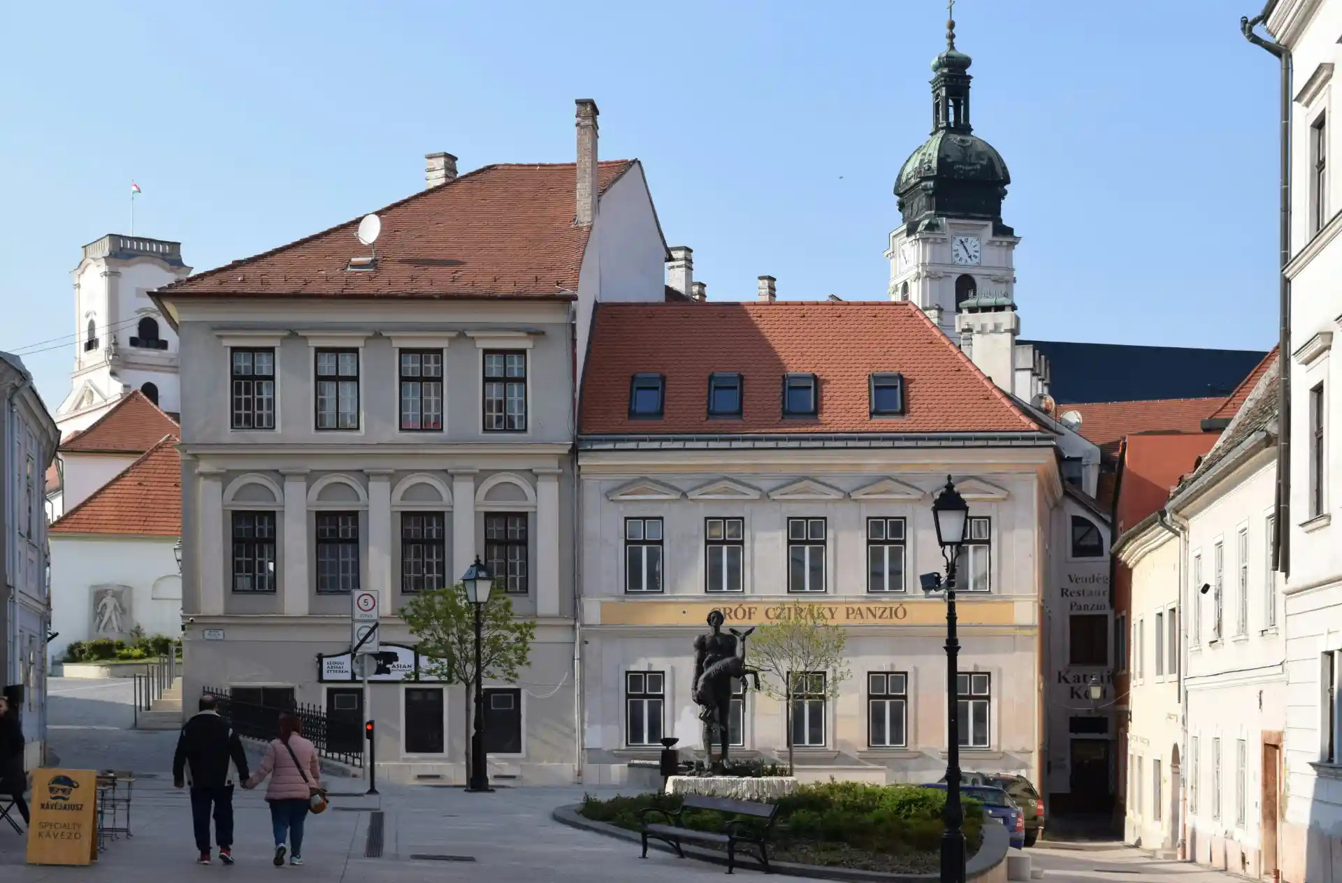 Historická budova s červenou strechou, vežou a sochou na námestí pod jasnou oblohou.