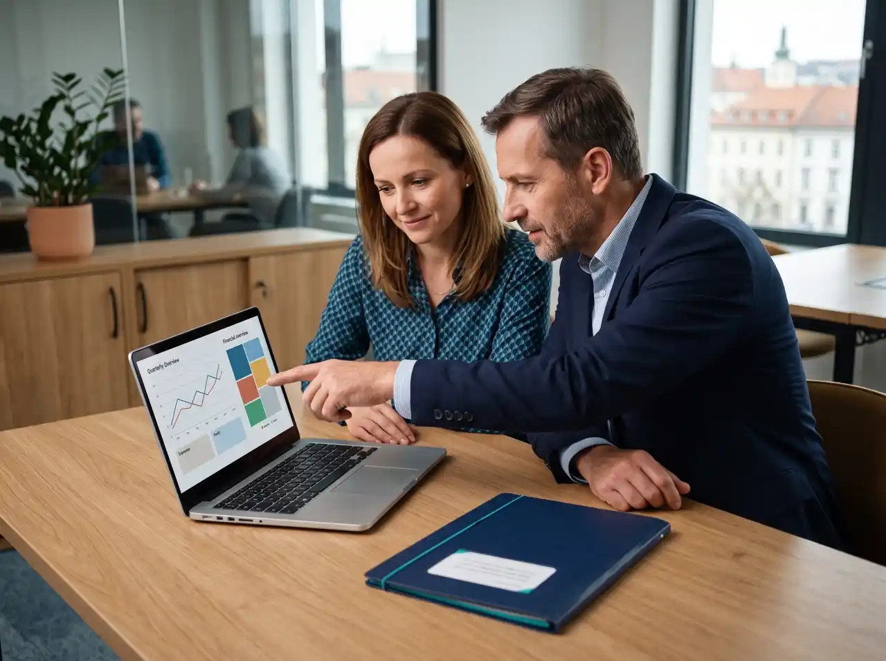 A man and a woman in an office are looking at charts on a laptop together, discussing business figures.